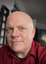 A bald white man with blue eyes looks at the camera with a very slight smile. He is wearing a maroon button-down shirt open at the collar, with a black T-shirt beneath. Behind him is a high-ceilinged room with knickknacks on the walls.